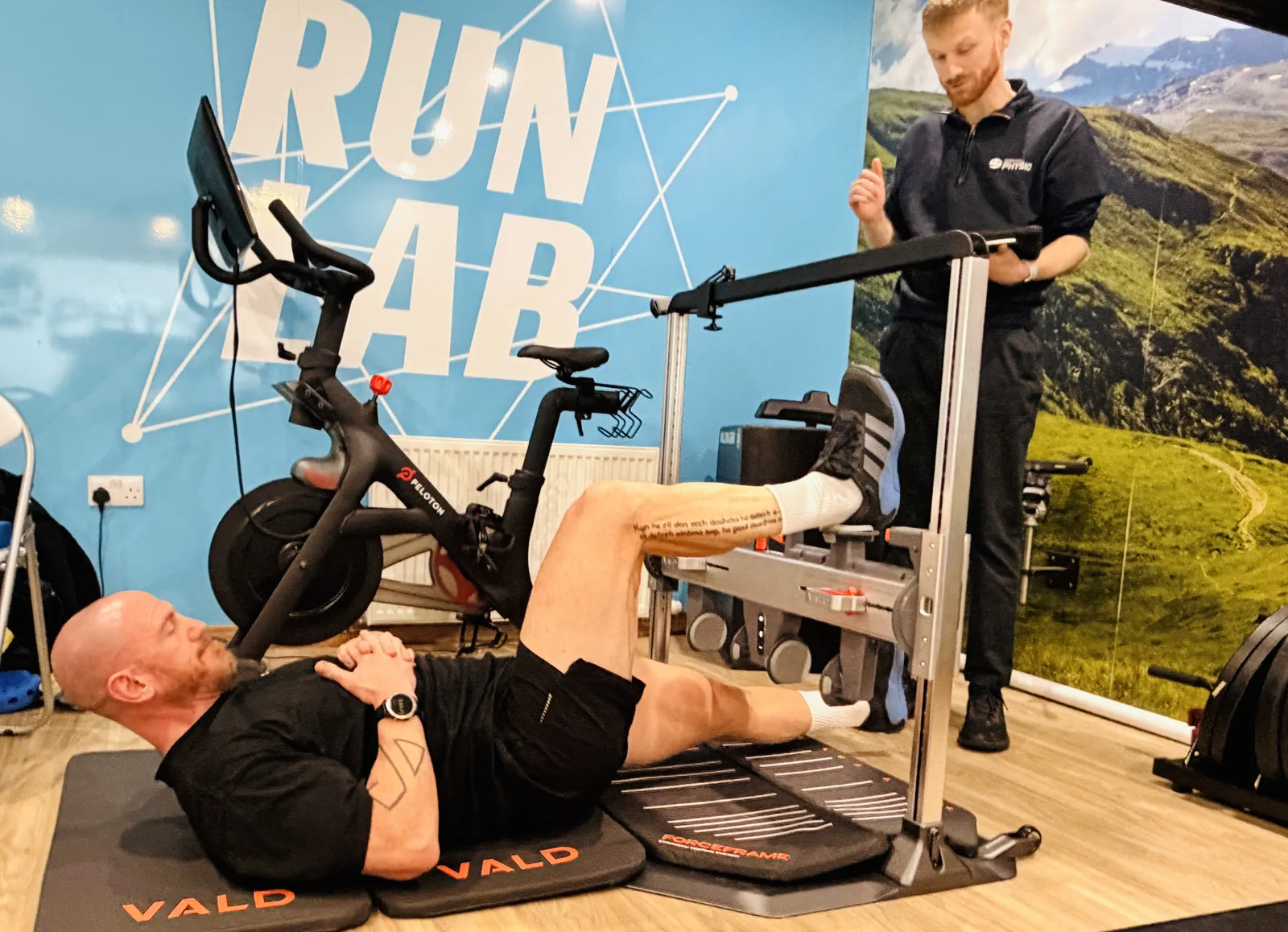 An athlete lying on the floor of The Run Lab® pushing one leg against the VALD® ForceFrame while a therapist reads the force on a tablet